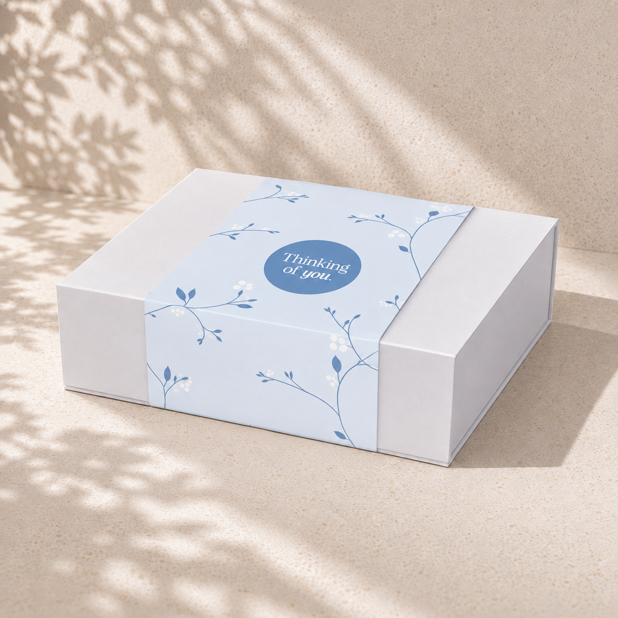 White magnetic close gift box with a blue Thinking of You botanical sleeve on a sunlit beige surface