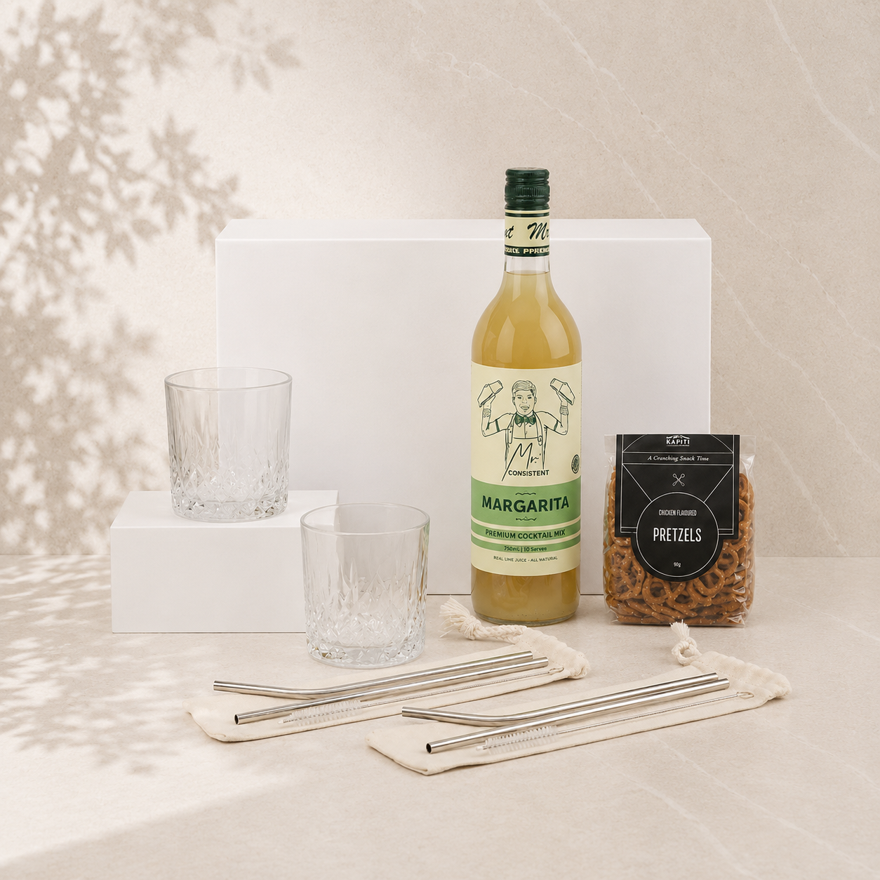 Cocktail gift hamper flat lay showing Mr. Consistent margarita mix, two crystal tumblers, Chocamama pretzels and mojito straw sets with white gift box