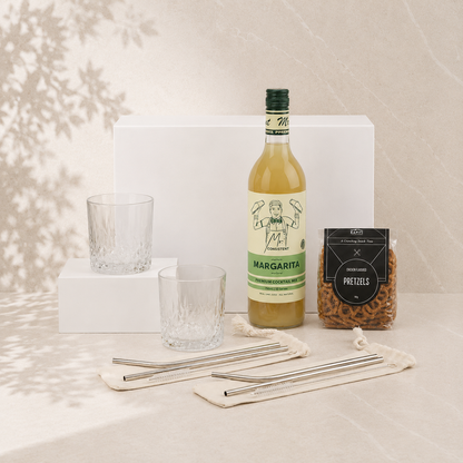 Cocktail gift hamper flat lay showing Mr. Consistent margarita mix, two crystal tumblers, Chocamama pretzels and mojito straw sets with white gift box