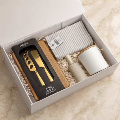 Settle In gift box open to show the acacia serving board, gold cheese knife set, vanilla candle, linen mist and tea towel packed in a white magnetic gift box