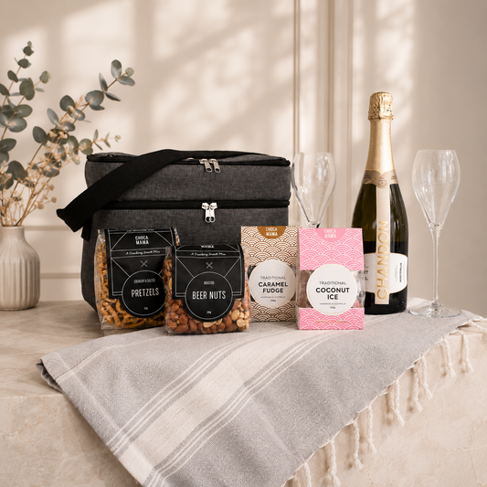 Luxury celebration gift hamper flat lay – Chandon sparkling wine, Krosno crystal champagne flutes, Chocamama pretzels, beer nuts, caramel fudge and coconut ice, presented in a grey RPET cooler bag on a Turkish towel