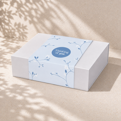 White magnetic close gift box with a blue Thinking of You botanical sleeve on a sunlit beige surface