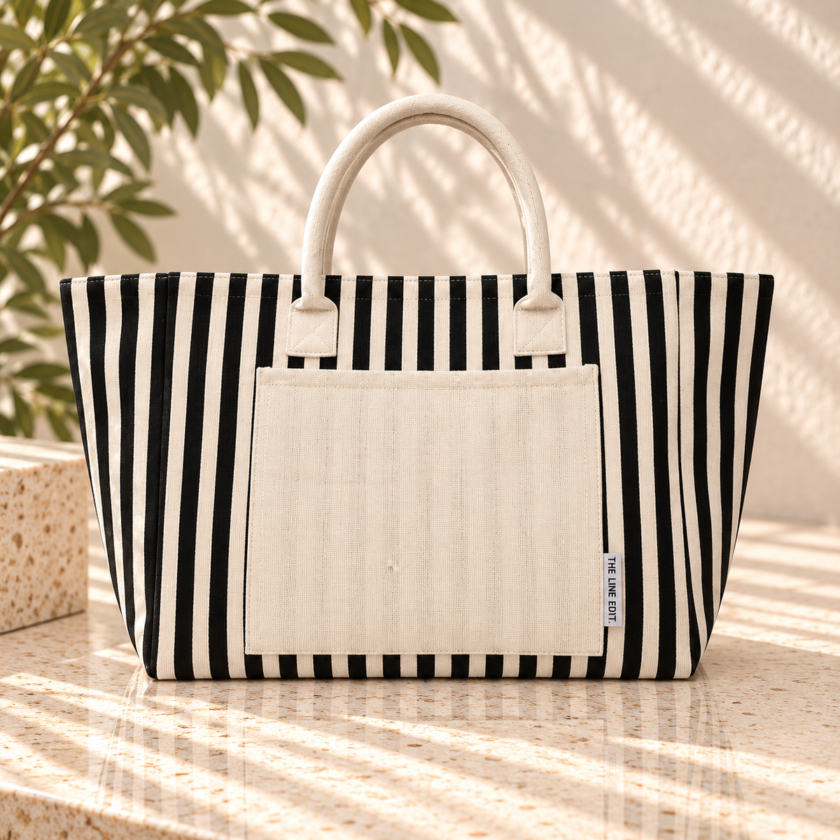 The Line Edit Hampton tote in black stripe with a cream canvas front pocket, standing in soft afternoon light