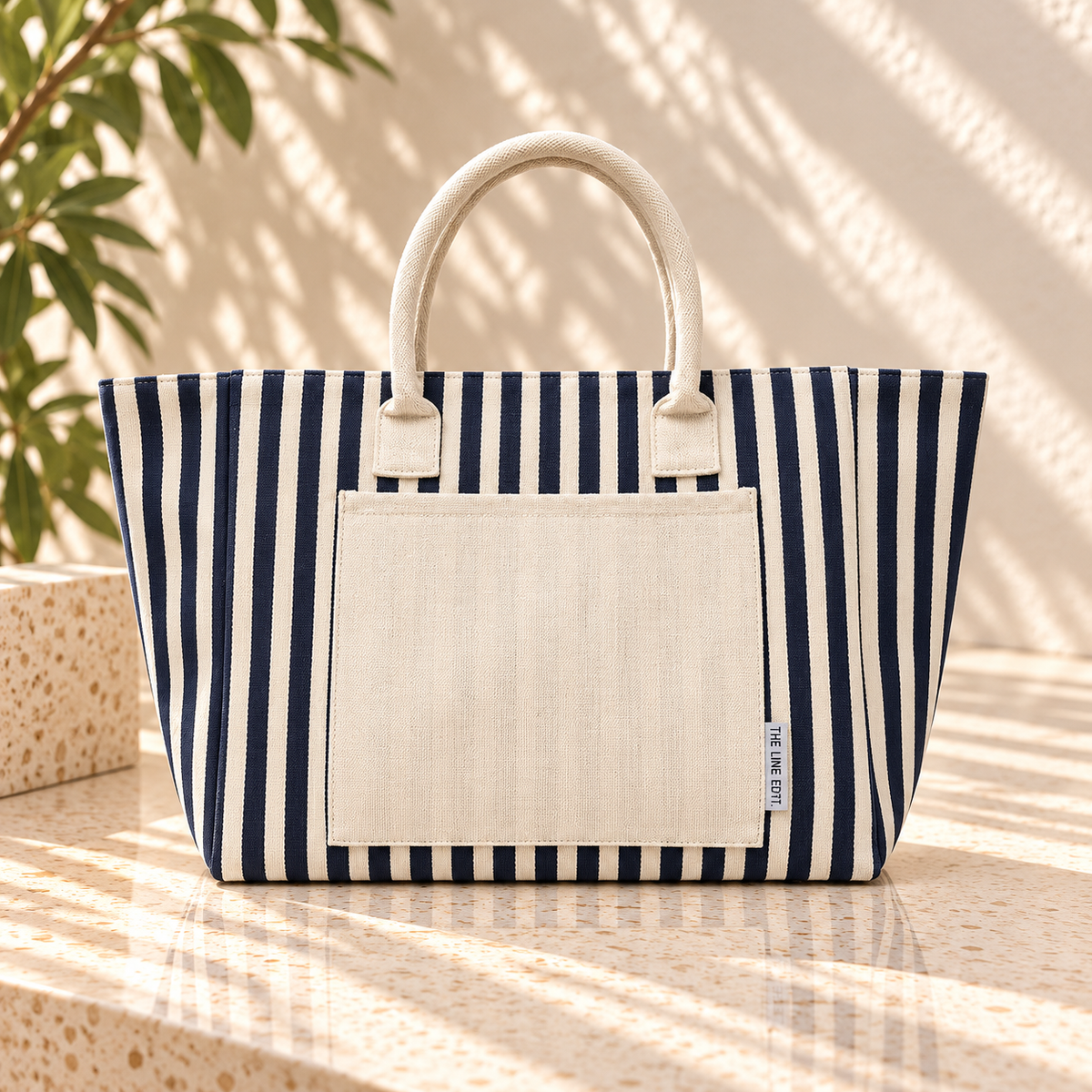 The Line Edit Hampton tote in navy stripe with a cream canvas front pocket, standing in soft afternoon light